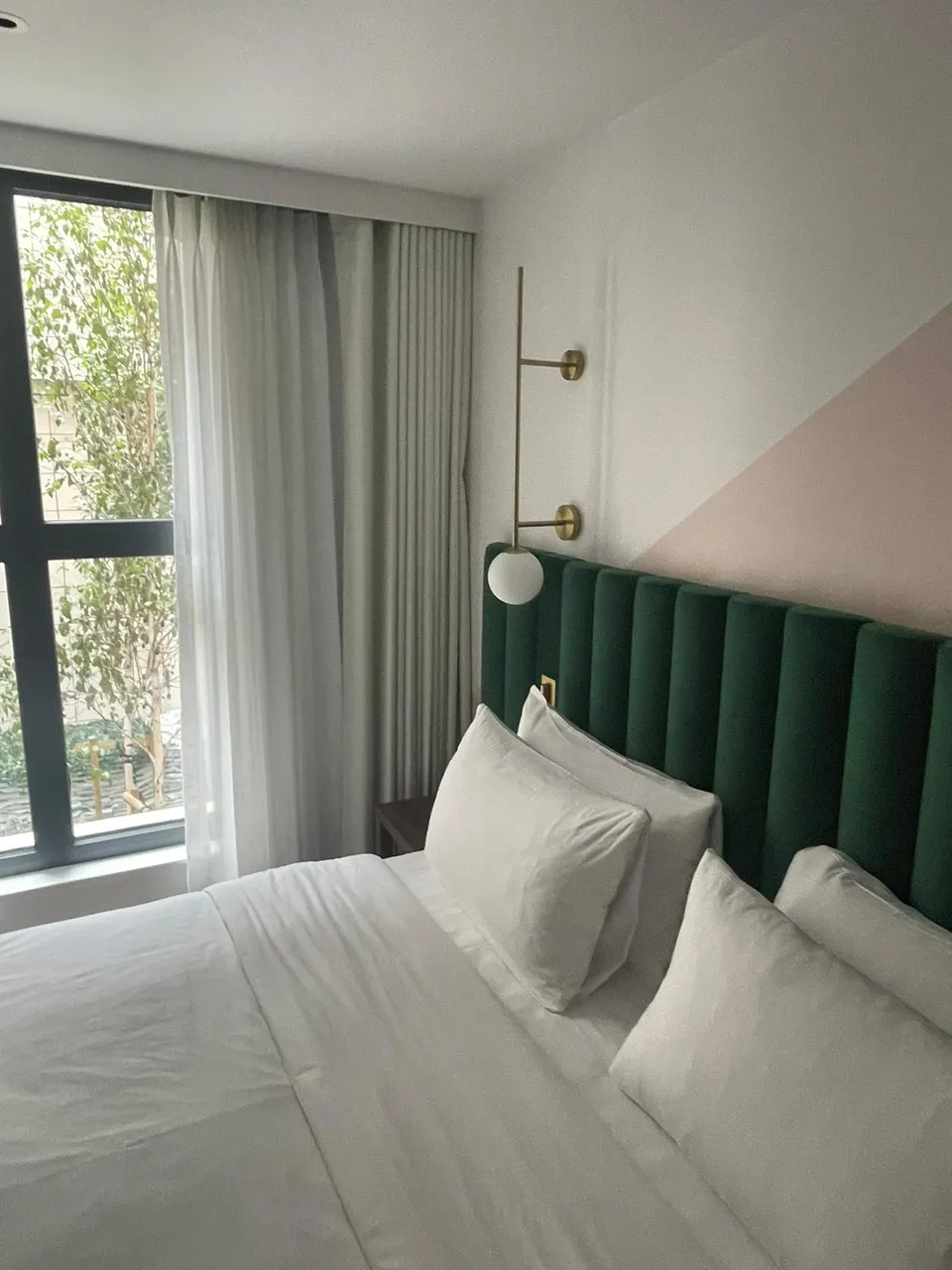 Grey curtain and white sheer at a tall window beside a green padded headboard in a hotel bedroom