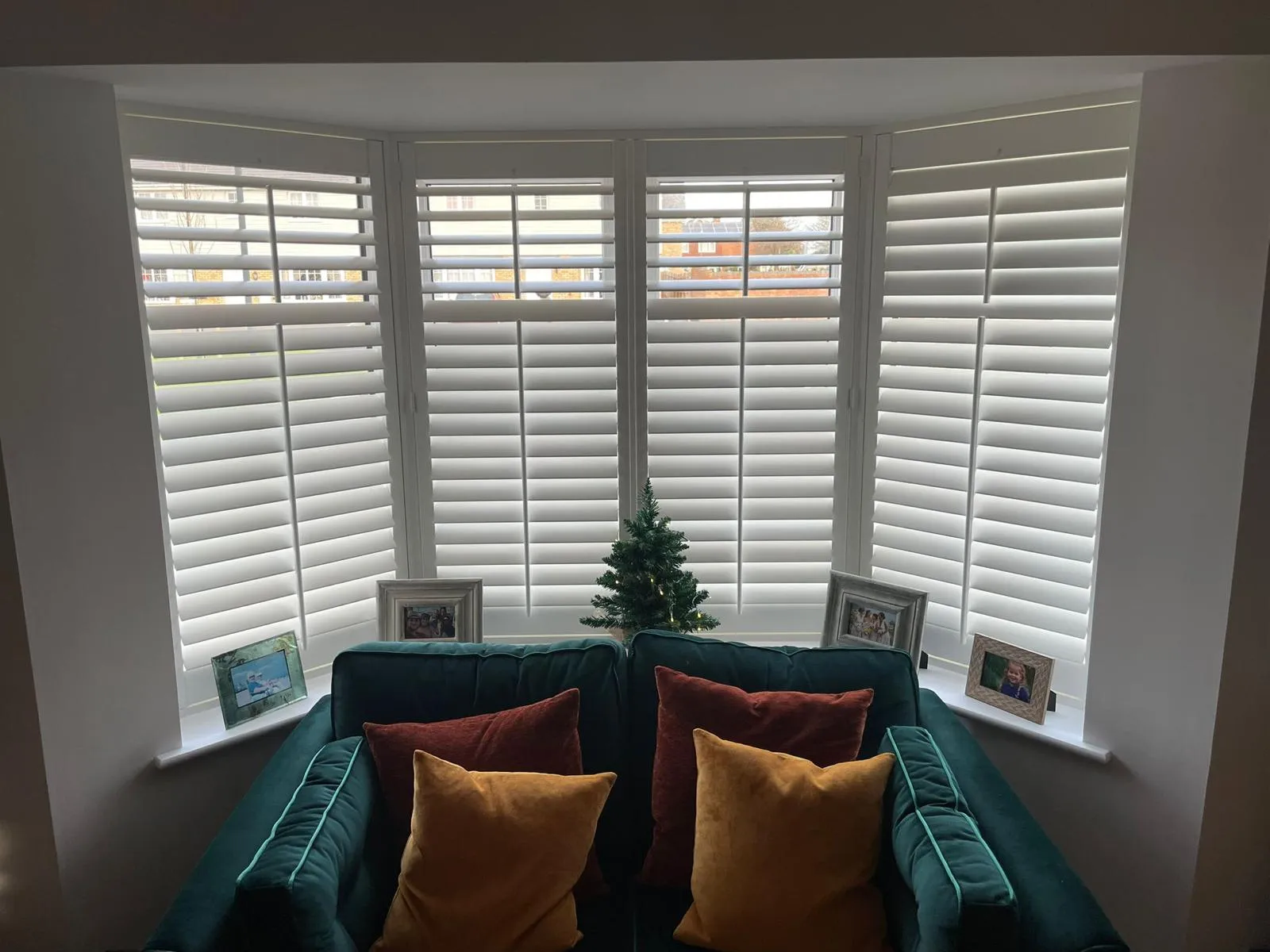 White plantation shutters across a bay window in a living room, with a teal sofa beneath