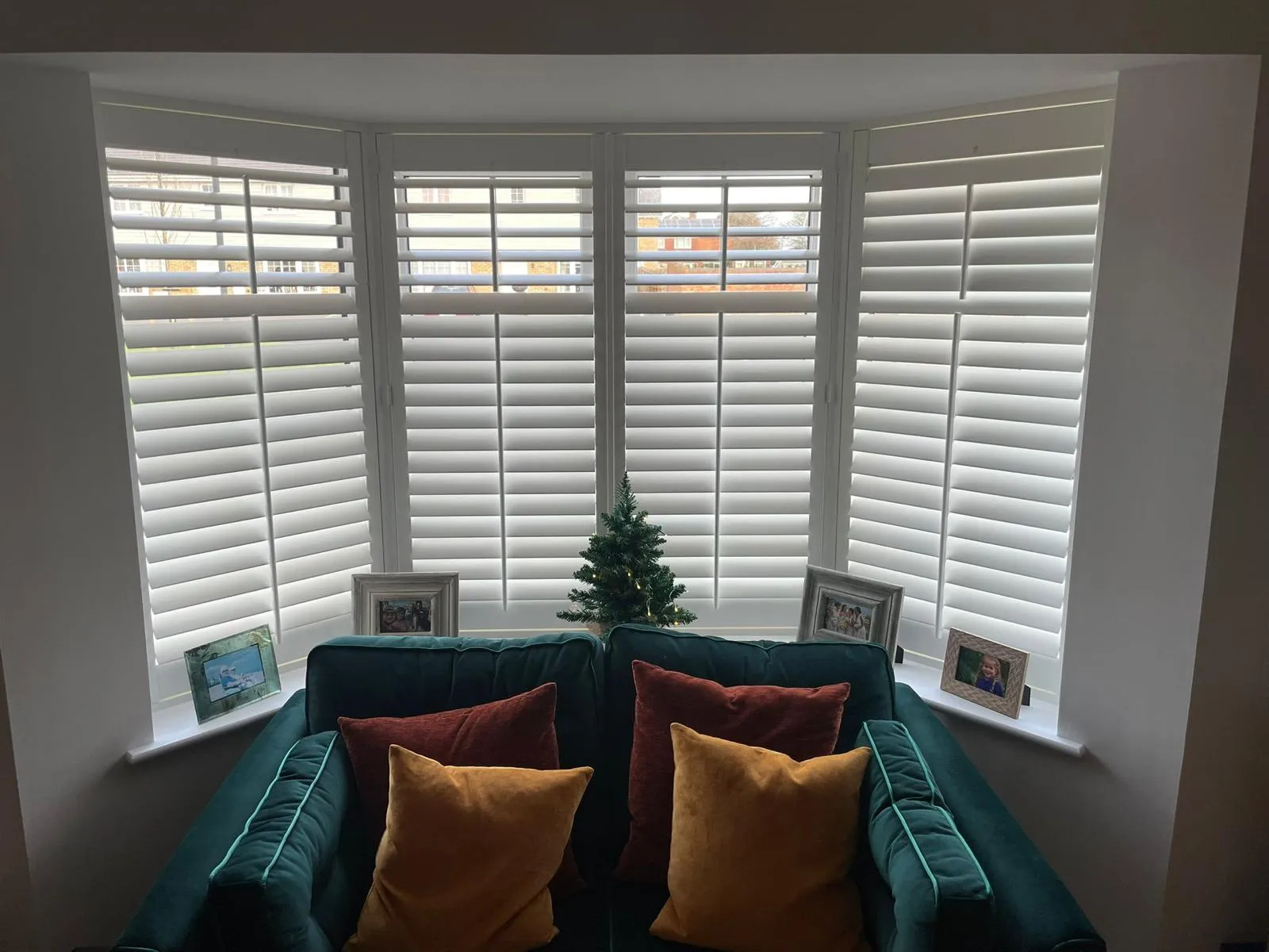 White plantation shutters in a bay window behind a teal sofa with orange and yellow cushions
