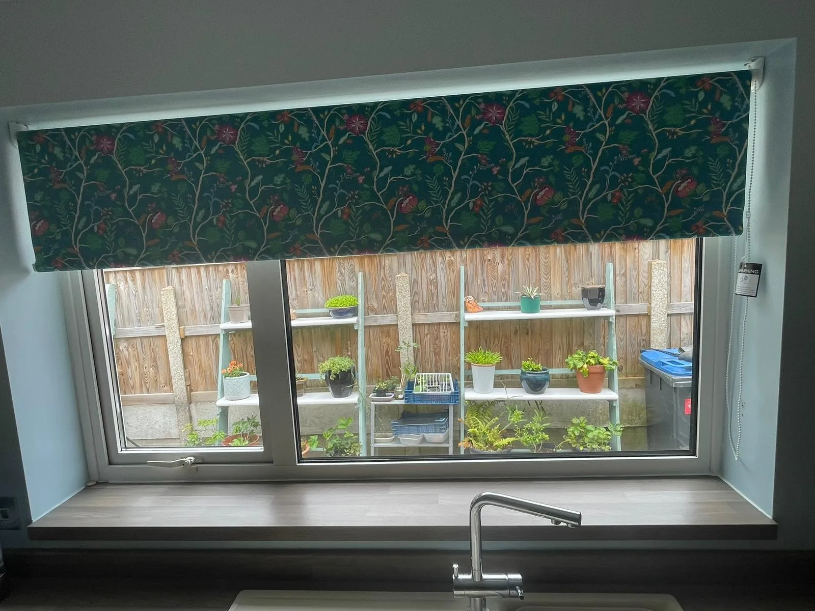 Dark green floral roller blind fitted above a kitchen sink window, with a garden view beyond