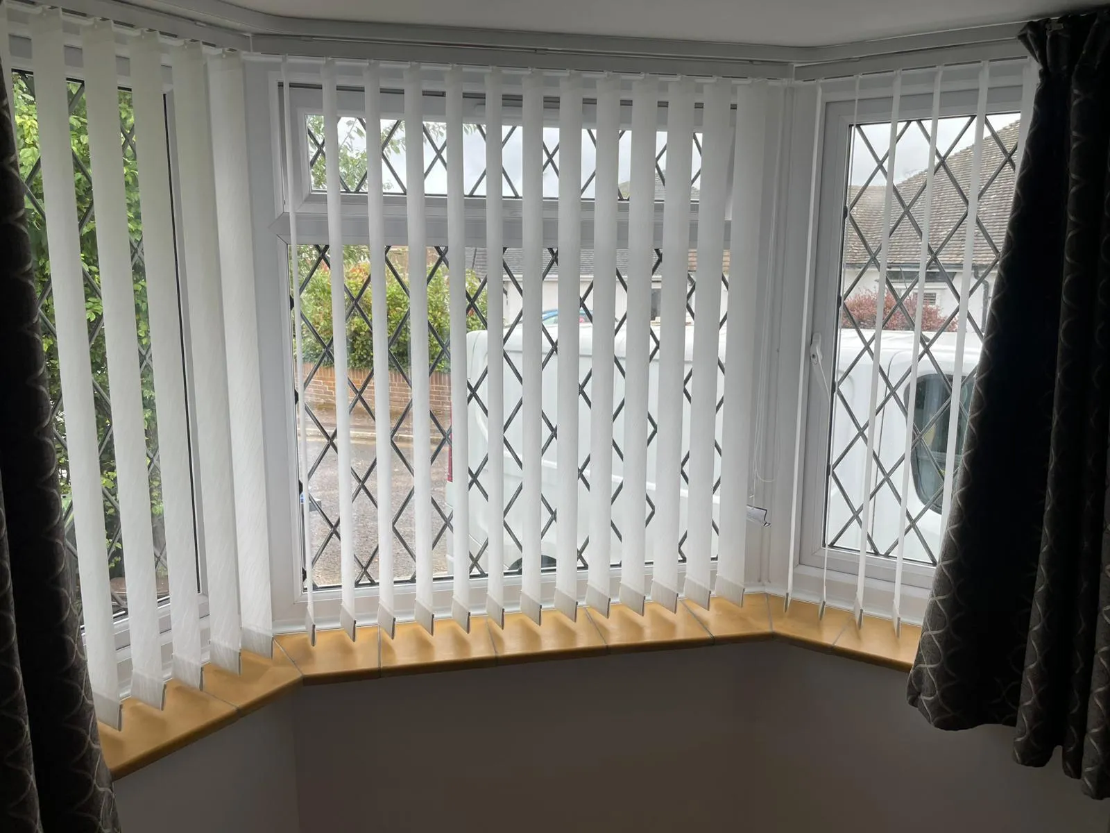 White vertical blinds and dark patterned curtains in a bay window with a wooden sill