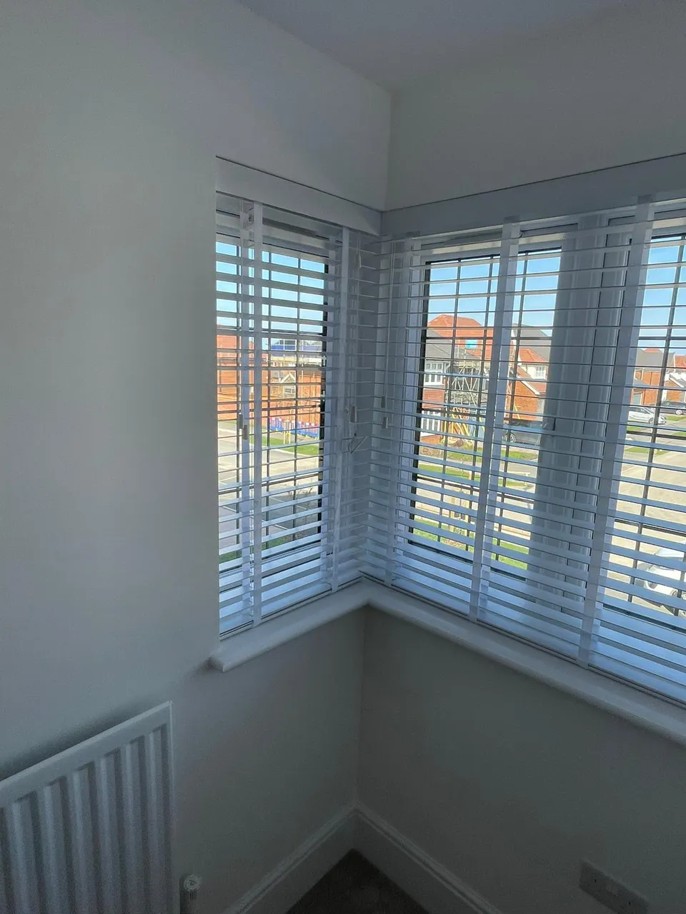 White venetian blinds fitted to a corner window in a bedroom, with rooftops beyond