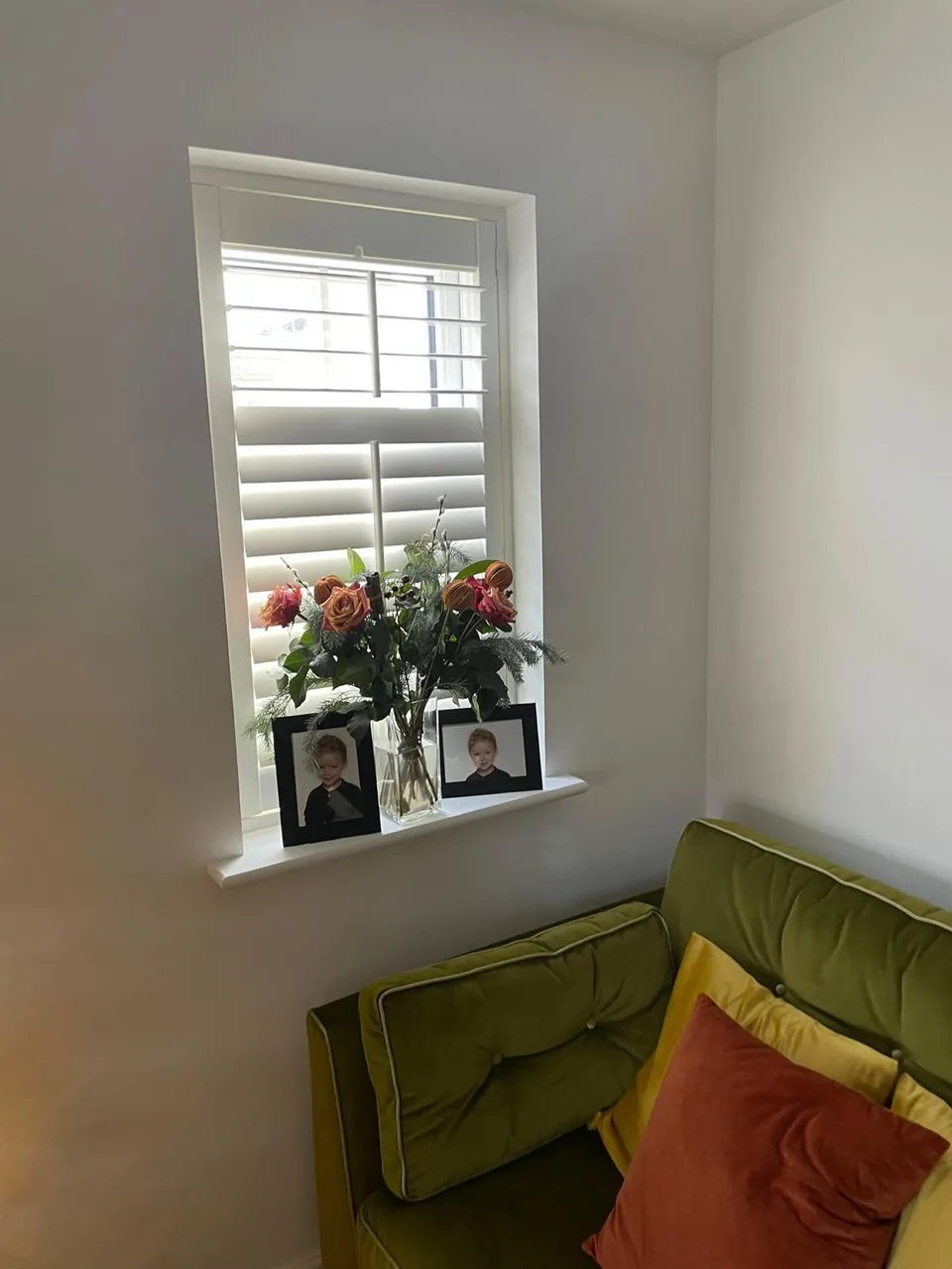 White plantation shutters on a small window above an olive green sofa in a living room